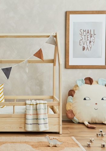 Creative composition of cozy scandinavian child's room interior with wooden bed, pillows, plush and wooden toys and textile hanging decorations. Neutral creative wall, carpet on the floor. Template.